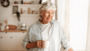 senior woman holding a cup in the kitchen standing next to a window looking out