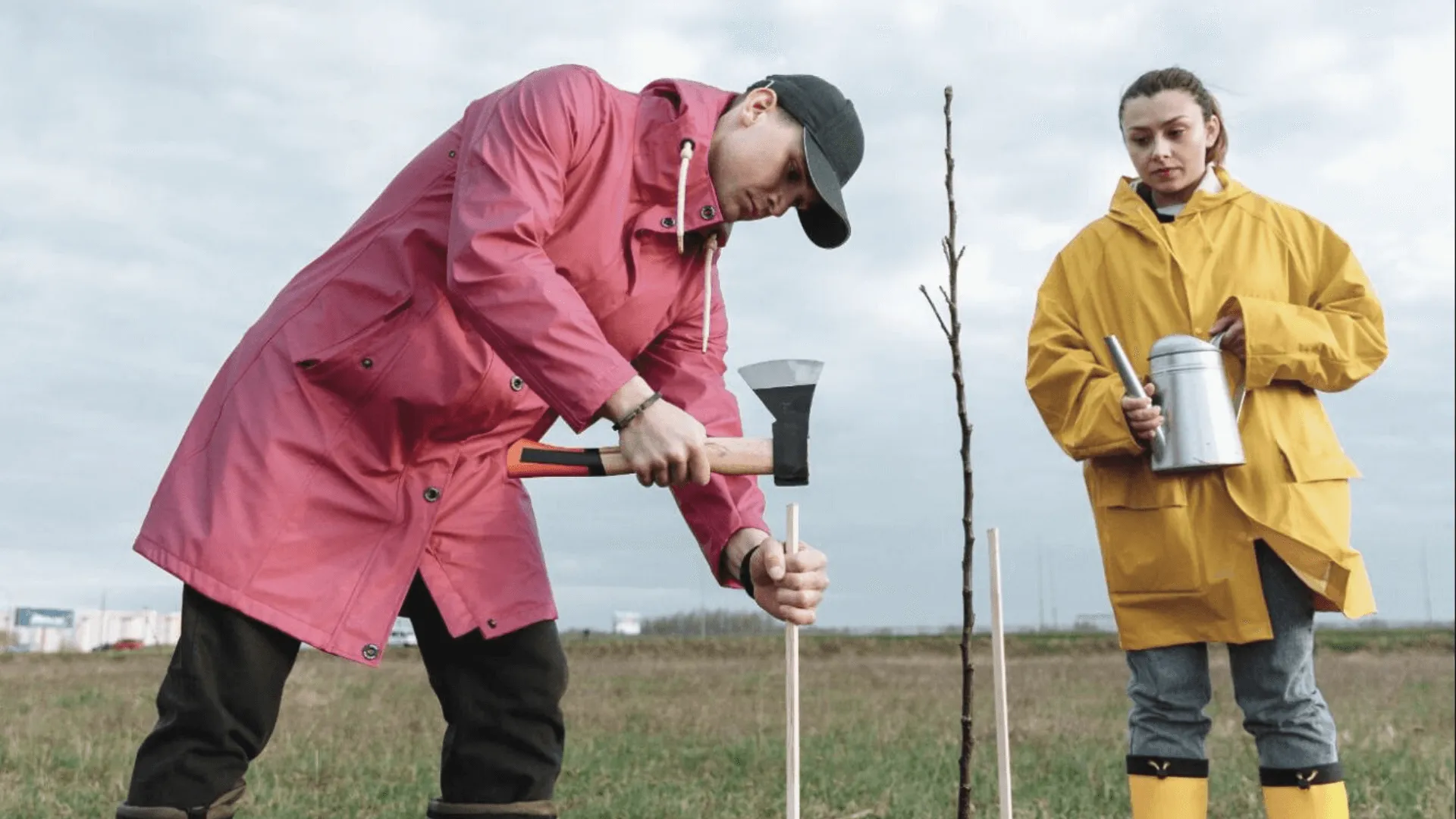 man in a red raincoat planting a tree while holding an axe with a woman in a yellow raincoat standing behind him, holding a watering can
