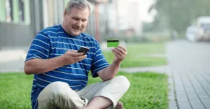 man sitting cross-legged on a lawn holding a phone and a credit card