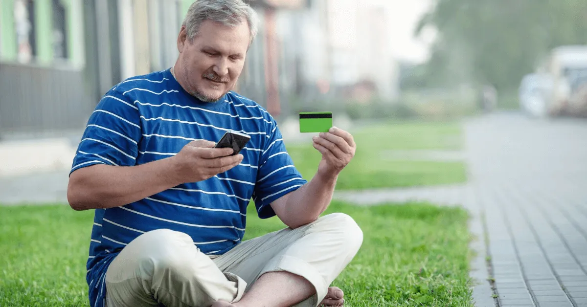 man sitting cross-legged on a lawn holding a phone and a credit card