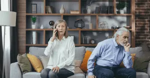 senior married couple sitting on a couch. The older woman is talking on the phone, and the older man is pensive