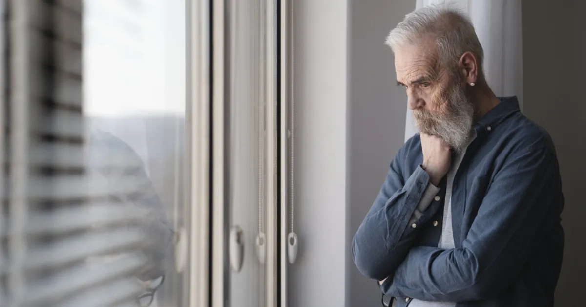 sad senior man looking out a window with blinds